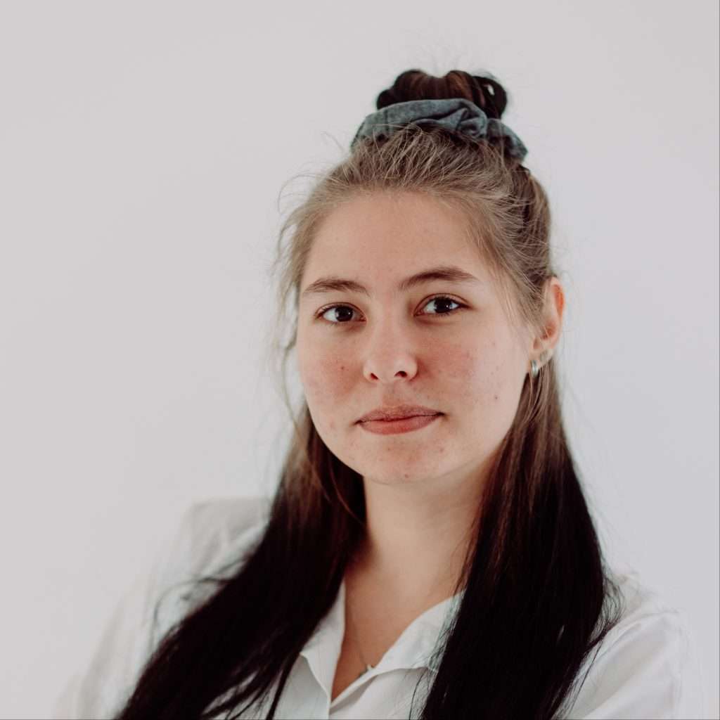 Close-up of a young woman's face from the nose up, with long dark hair and a grey scrunchie in a top knot, wearing a light shirt against a pale background.
